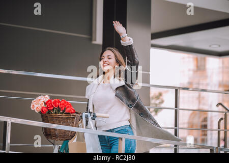 Bella giovane donna sorridente camminando con la bicicletta e agitando la mano sulla strada della citta'. Bellezza,gesto e concetto di stile di vita Foto Stock