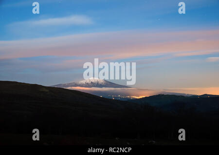 Il monte Etna ricoperta di neve con nebbia e nuvole al tramonto. Il cono di il più alto vulcano attivo in Europa di notte. Vista da Cesarò, parco dei Nebrodi Foto Stock