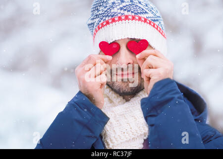 Giovane uomo caucasico indossando giacca blu, cappello e sciarpa tenendo due cuori rossi davanti ai suoi occhi all'aperto per il giorno di San Valentino Foto Stock