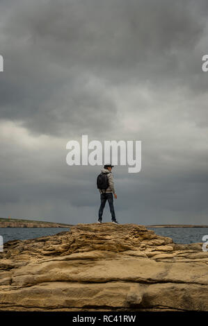 Uomo con zaino in piedi sulla parte superiore delle rocce che guarda al mare, moody sky. Foto Stock