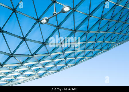 Architettura moderna tetto di vetro con design curvo della struttura in acciaio. Basso angolo vista con cielo blu, illuminazione interna e parete bianca dell'edificio Foto Stock