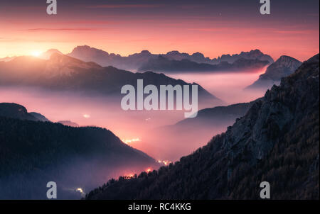 Montagne nella nebbia al bel tramonto in autunno nelle Dolomiti, Italia. Paesaggio alpino con mountain valley, nuvole basse, foresta, viola sky city sistema luminoso Foto Stock