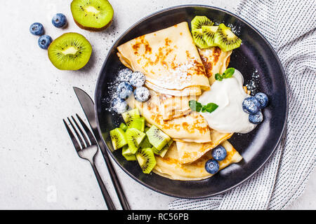 Sottile crepes con crema di ricotta, kiwi e mirtilli in una piastra nera. Cucina russa concetto. Foto Stock