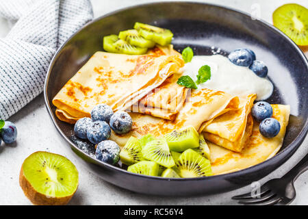 Sottile crepes con crema di ricotta, kiwi e mirtilli in una piastra nera. Cucina russa concetto. Foto Stock