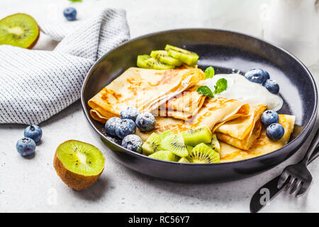 Sottile crepes con crema di ricotta, kiwi e mirtilli in una piastra nera. Cucina russa concetto. Foto Stock