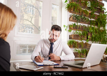 Colpo di vendite uomo seduto alla scrivania in ufficio e facendo alcuni documenti cartacei durante la consultazione con il suo collega. Il lavoro di squadra in ufficio. Foto Stock