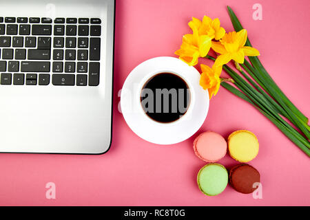 Donna o area di lavoro femminile con laptop, notebook, tazza di caffè, macarons, e fiori di narciso rosa sul tavolo. Vista dall'alto. Lay piatto. Copia dello spazio. Foto Stock