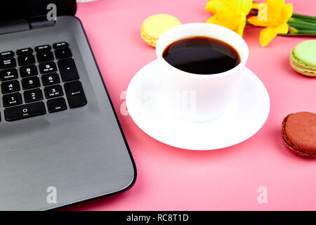 Donna o area di lavoro femminile con laptop, notebook, tazza di caffè, macarons, e fiori di narciso rosa sul tavolo. Vista dall'alto. Lay piatto. Copia dello spazio. Foto Stock