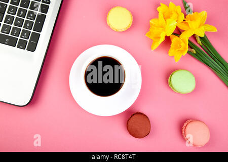 Donna o area di lavoro femminile con laptop, notebook, tazza di caffè, macarons, e fiori di narciso rosa sul tavolo. Vista dall'alto. Lay piatto. Copia dello spazio. Foto Stock