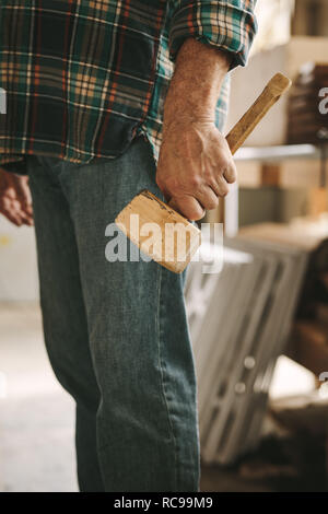 Ritagliato colpo di falegname tenendo un mazzuolo di legno martello in officina. Uomo con un mazzuolo martello in mano al laboratorio di falegnameria. Foto Stock