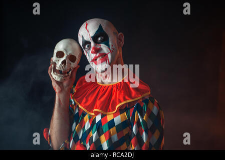 Bloody clown con crazy face detiene il teschio umano, terribile segreto. L'uomo con il trucco in costume di carnevale, mad maniac Foto Stock