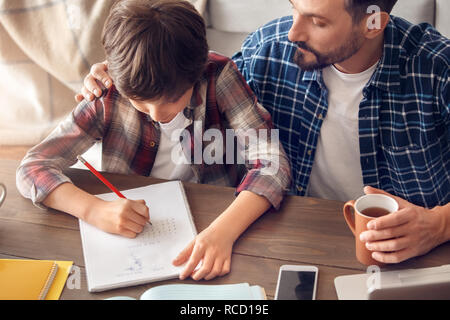 Il padre e il figlio insieme a casa seduti a tavola il papà di bere il tè caldo guardando boy facendo esercizio di matematica toccando il suo sostegno houlder Foto Stock