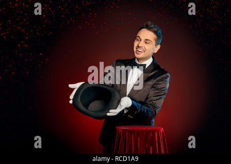 Professional mago indossare tuta e guanti isolati permanente su nero e sfondo rosso rendendo trucco mostra vuoto top hat al pubblico sorridente ven Foto Stock