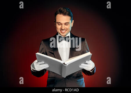 Professional mago tuta da indossare guanti di nd in piedi isolato su nero e sfondo rosso azienda libro magico incantesimo lettura sorridente allegro Foto Stock