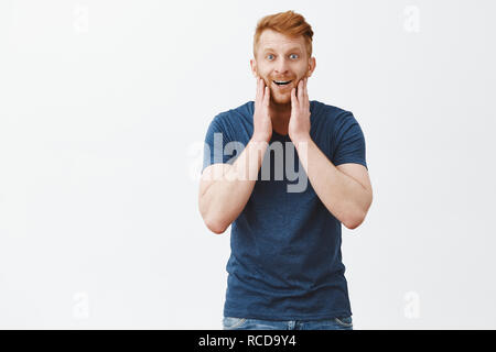 Soddisfatto e stupito guy guardando in uno specchio, soddisfatto con un nuovo taglio di capelli. Bell'aspetto maschile e bello redhead maschio modello in blu t-shirt, toccando le guance e guardando con un ampio sorriso alla fotocamera Foto Stock