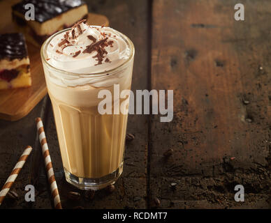 Vetro di elevata di cappuccino schiumoso caffè con topping di cioccolato servito con torta e caramelle sul vecchio legno scuro tavolo. Spazio di copia Foto Stock