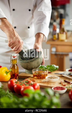 Lo Chef macinazione di erbe aromatiche fresche in un pestello e mortaio durante la preparazione di un pasto con freschi ingredienti sani in un vicino fino alle mani Foto Stock