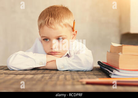 Pupilla pigro. Triste piccolo scolaro seduti a tavola e che poggia il mento sulle mani piegate, di non voler fare un sacco di compiti Foto Stock