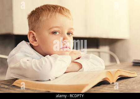 Antipatia per la lettura. Triste piccolo ragazzo seduto al tavolo di appoggio e il mento sulle mani ripiegato su un libro aperto, essendo stanchi per la lettura di un libro Foto Stock