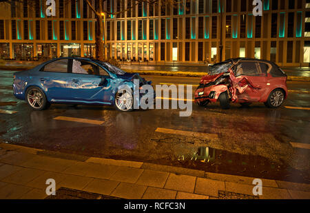 Due auto incidente. Si è schiantato vetture su strada sulla posizione della città di notte. Una berlina blu contro una città rossa auto. Collisione frontale con grossi danni. Foto Stock