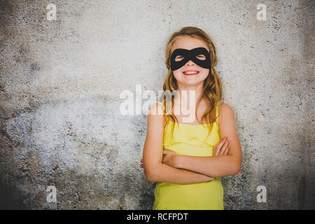 Ragazza bionda con nero maschera di supereroi e maglietta gialla è in posa di fronte di sfondo in calcestruzzo Foto Stock