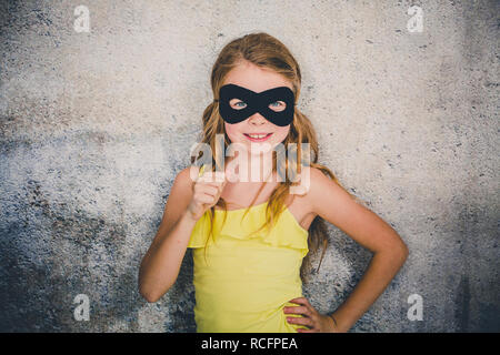 Ragazza bionda con nero maschera di supereroi e maglietta gialla è in posa di fronte di sfondo in calcestruzzo Foto Stock