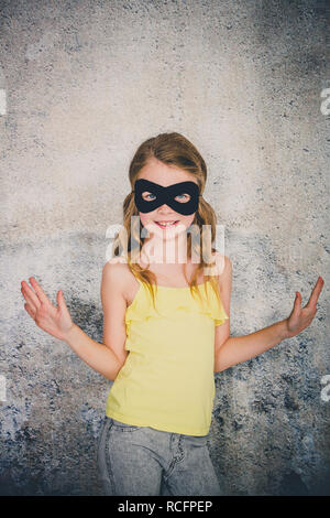 Ragazza bionda con nero maschera di supereroi e maglietta gialla è in posa di fronte di sfondo in calcestruzzo Foto Stock