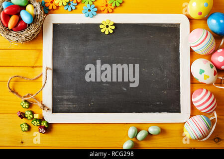 Layout di pasqua su giallo di tavoli in legno di uova colorate e caramelle intorno alla lavagna vuota tablet in cornice di legno per spazio di copia Foto Stock
