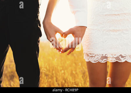 Vista posteriore del giovane in amore cuore di gesti con le dita. Close up di un uomo e di una donna che fa il cuore con le mani contro il sole splendente su golden erba ba Foto Stock