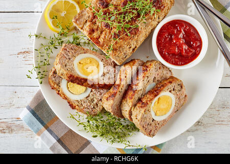 Fette di polpettone farcito con uova sode servite con salsa di pomodoro e le fette di limone su una piastra bianca con la forchetta e coltello su un vecchio tavolo di legno, v Foto Stock