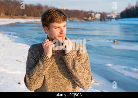 Uomo barba lunga bello hipster in maglieria maglione alto colletto stand frozen river sfondo. Paesaggio invernale bellezza. L'uomo godere l'inverno in un giorno di sole. In inverno abiti di maglia in fase di riscaldamento. Inverno moda. Foto Stock