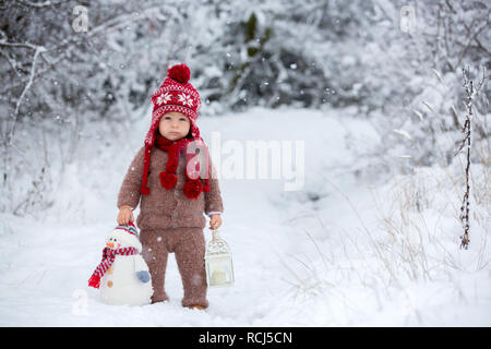 Ritratto di un simpatico bimbo baby vestito con una mano di colore marrone camicia a maglia, pantaloni, Red Hat e sciarpa, tenendo lanterna e pupazzo di neve, passeggiate attraverso la snowy Foto Stock