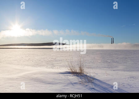 Panorama di un lago ghiacciato con comignoli fumanti nella nebbia, il sole e il sentiero dal piano. Foto Stock