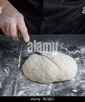 Cuocere in uniforme nero taglia un pezzo rotondo di pasta impastata con un coltello su un tavolo di legno Foto Stock