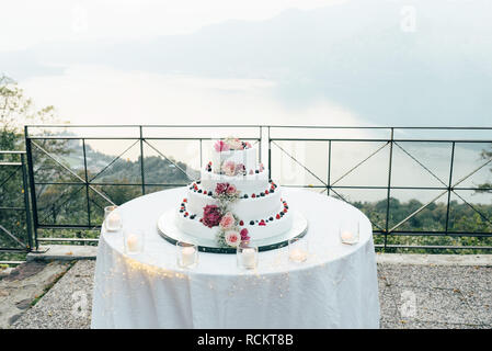 Una bella torta di nozze in quattro livelli è sul tavolo contro lo sfondo del pittoresco lago Foto Stock