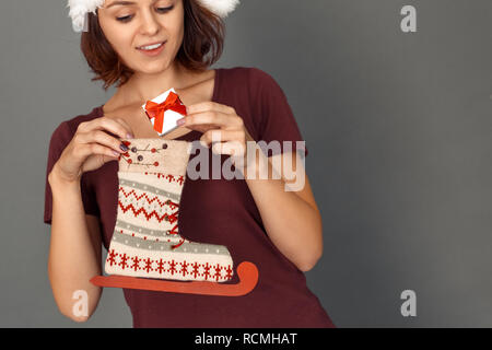 Giovane donna indossa santa hat tema natalizio isolato in piedi sul muro grigio messa piccola scatola regalo decorata in skate boot sorridendo eccitato close-up Foto Stock