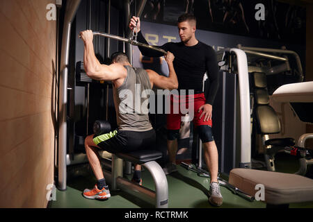 Due giovani uomini, 20-29 anni. Un uomo esercizio di muscoli della schiena, formazione sul lat pulldown di macchina, mentre altri amico o un allenatore è aiutare lui. In palestra, wit Foto Stock