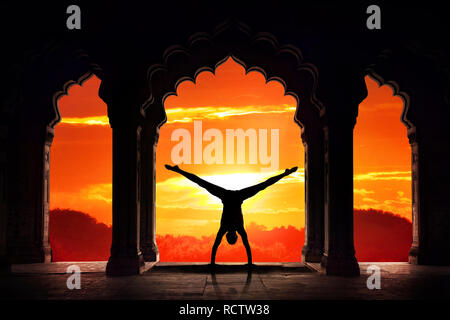 Silhouette uomo fare yoga anticipo handstand asana nel vecchio tempio in arancione tramonto sullo sfondo del cielo Foto Stock