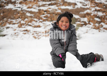 Latino sorridente ragazza seduta sulla coperta di neve montagna, facendo un pupazzo di neve e di indossare abbigliamento invernale. Foto Stock