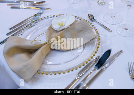 Raffinati ristoranti impostazione tabella con lastre trasparenti, beige tovagliolo di lino naturale con orchid e decorazioni dorate pronto per gli ospiti di un matrimonio Foto Stock