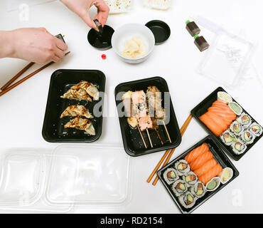 Vista da sopra della tabella contenente il sushi casella consegna con sushi california, Maki, sashimi, yakitori e bastoni di legno con wasabi e salsa di soia - donna preparare la salsa di soia Foto Stock