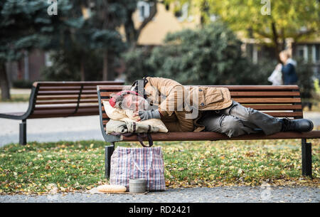 Senzatetto mendicante uomo con un sacco giacente sul banco di prova all'aperto in città, dormendo. Foto Stock