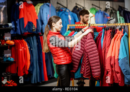 Giovane donna scelta di abbigliamento invernale picking up down jacket sulla bilancella nel negozio di articoli sportivi Foto Stock