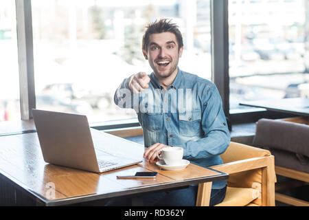 Hey! Ritratto di felice positivo giovane barbuto freelancer in blue jeans shirt sono seduti in cafe e di lavoro sul computer portatile con sorriso toothy e punto Foto Stock