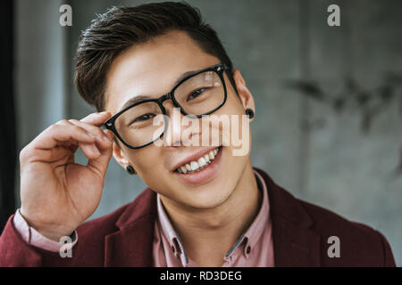 Ritratto di bel giovane uomo asiatico la regolazione degli occhiali e sorridente in telecamera Foto Stock
