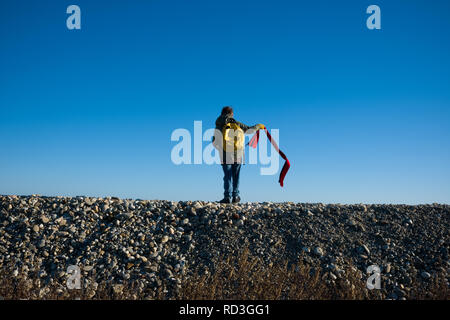 Parte posteriore del ragazzo di 11 anni che porta uno zaino e oscillando la sua sciarpa rossa, sorge su di un ciottolo causeway affacciata sul mare incorniciato da un cielo blu chiaro Foto Stock