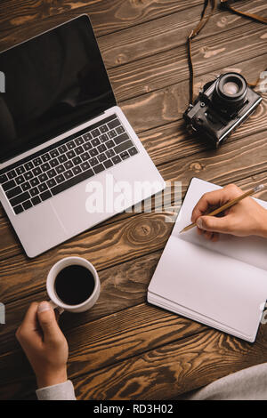 Vista ritagliata del fotografo maschio iscritto in notebook a tavola con la tazza di caffè, laptop e retrò fotocamera foto Foto Stock