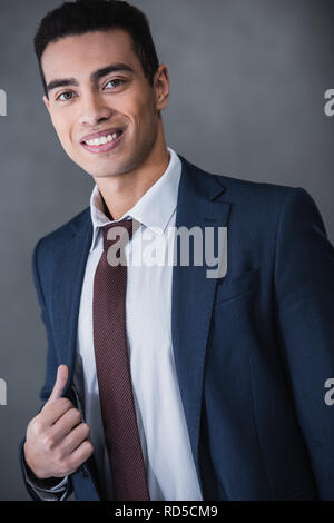 Ritratto di bel giovane razza mista imprenditore tuta regolazione camicia e sorridente in telecamera su grigio Foto Stock