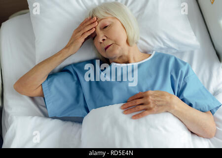 Sconvolto senior donna giaceva nel letto di ospedale, toccando la fronte e aver mal di testa Foto Stock