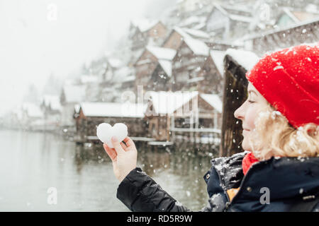 Bionda e giovane donna sorridente in un Red Hat tenendo palla di neve in una forma di un cuore di fronte al vecchio villaggio di Hallstatt, Austria. Sfondo sfocato, Foto Stock